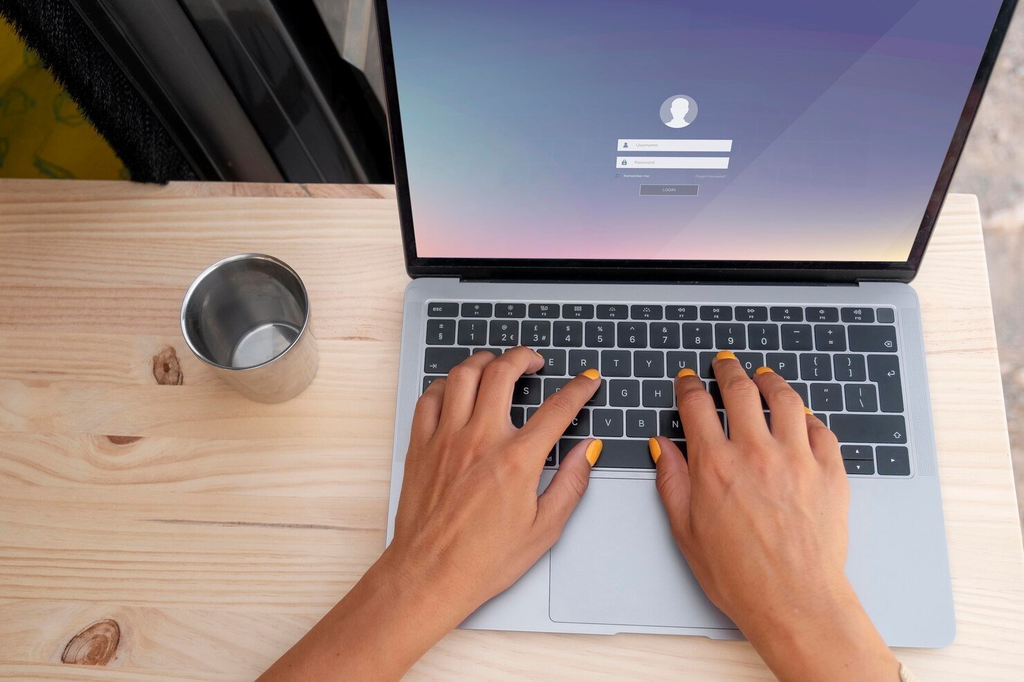 Login on PC Hands with yellow-painted nails type on a laptop with a login screen displayed. A metal cup sits beside it on a wooden desk.