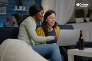 Two women happily looking at a laptop together on a comfortable blue couch. A beer bottle is on a white coffee table, and a friend in the background holds a red cup.