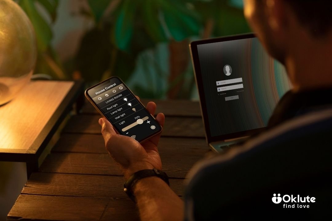 A person holds a smartphone showing a home control app with light settings in a dimly lit room. A laptop with a login screen is in the background.